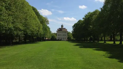Philip Cornwall A large three-storey  country house as viewed from the end of a large lawn, rows of trees either side.