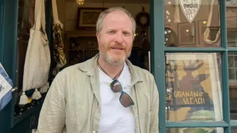 Pauric Lawne is stadning in a shop doorway, wearing a white tshirt and beige coat. He has short blond hair and beard.