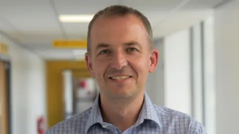 Nottingham University Hospitals NHS Trust A man in a light chequered shirt stood in a hospital corridor.