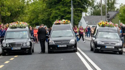 Pa Media three black Heares are sorted side by side on one road. Each one has a flower placed on it. There are a few people standing behind cars.