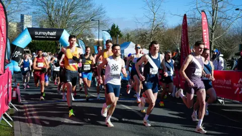 A group of runners sets off from a start line with an inflatable arch in the background and banners and fences along the edge of the road. The sun is shining