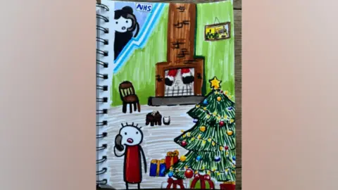 The winning Christmas card drawn in a notepad. The design shows a person on the phone inside a house with a Christmas tree, presents and two legs in a Santa costume dangling down the chimney. One corner of the page is framed off as an inset, to show a woman with long dark hair wearing a headset, with the NHS logo on the top. Inside the house is also a picture of an ambulance hanging on the wall and a cat in front of the chimney.