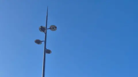 Plymouth City Council A large lighting column against a blue sky. The light is very tall and grey. It has four multi-directional bulbs.
