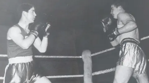 Paul Gilfeather Una fotografía antigua en blanco y negro de un joven Frank Gilfeather con pantalones cortos de boxeo y una camiseta sin mangas con la bandera de Escocia en el centro, en un combate de boxeo contra un hombre más alto.