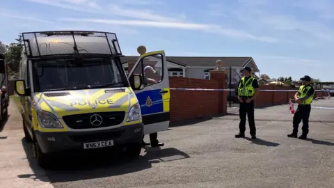 BBC Three police officers stand next to crime scene tape and a police van. Behind them, a tarmac area with red brick wall, forensics tent and small cones.
Behind the wall, the tops of small dwellings can be seen.