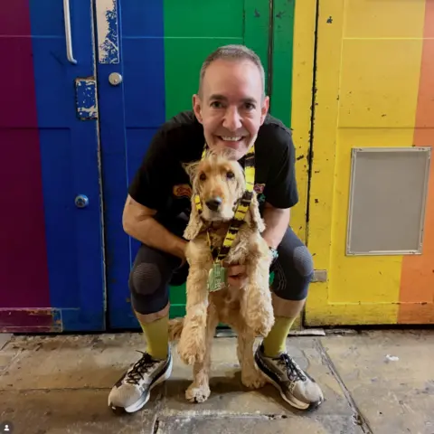 Heaven A man dressed in running gear and a big smile crouches with his dog against a rainbow wall.