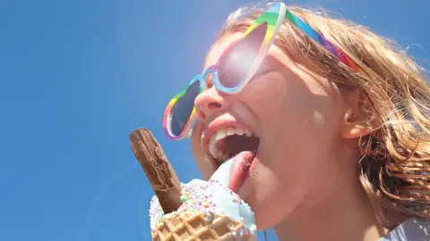 A girl with blonde hair and heart-shaped sunglasses licking an ice cream with a chocolate flake
