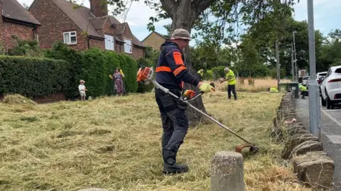 Handout A man wearing a camouflage bucket hat pictured trimming grass next to a pavement.