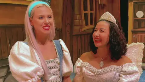 Kitty Harris, with long blonde hair and a bright blue headband, is dressed as Cinderella on stage alongside Hayley Tamaddon, with black curly hair, a white tiara and white winged fairy godmother costume