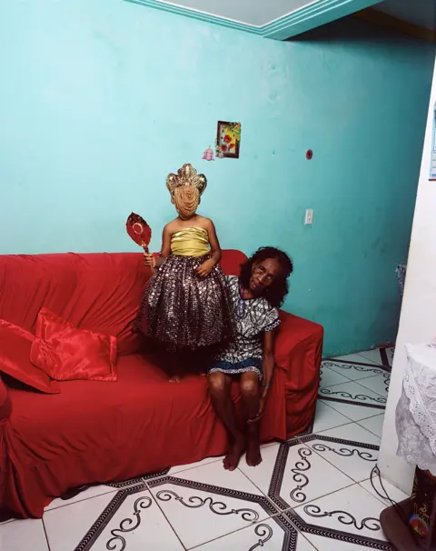 Deana Lawson A photo by Deana Lawson of a child wearing a headdress stood on a red sofa, next to a woman who is sitting down