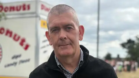 A man looking at the camera. He has short grey hair. She is wearing a black jacket and black and white checked shirt.
