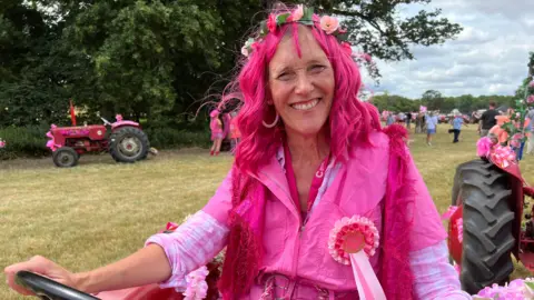 Mousumi Bakshi/BBC A woman sitting on a tractor. She wears a pink wing, floral headpiece, pink overalls and there is a pink rosette pinned to her chest. 
