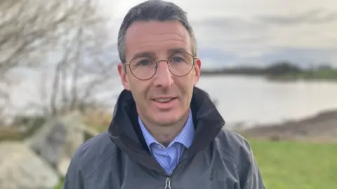 Andrew Muir stands outside. He wears a grey coat and blue shirt. He has glasses and grey hair. There is grass, water and trees behind him. 