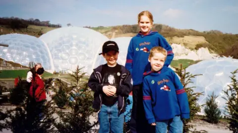 Rosie Johns and her two brothers are posing for the photo and behind them are the biomes. Rosie and one of her brothers are wearing Harry Potter branded hoodies and the other brother is wearing a cap. One of the brothers is pulling a funny face.