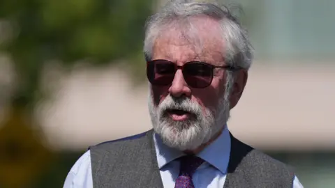 PA Media Gerry Adams is pictured from the chest up - he wears a blue shirt, grey waistcoat, dark sunglasses and purple tie