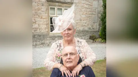 Sue Stollery A woman stands outside with her arms on a man's shoulders. They are both dressed in formal wedding attire