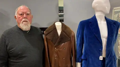 BBC A man in a grey jumper standing next to two items of clothing. He has a grey beard and grey hair. Next to him is a brown leather jacket and a blue velvet suit which are on mannequins.