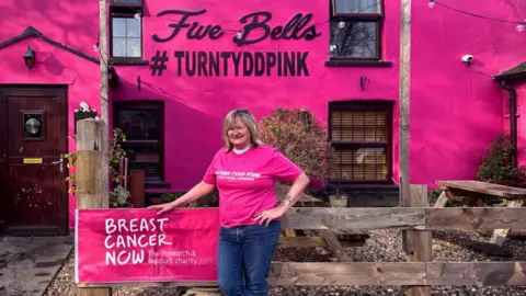 Supplied A woman with shoulder-length blonde hair standing in front of a pink pub with the words "Five Bells" and "#turntyddpink" in black. She is wearing a pink top and blue jeans, and is smiling and resting her hand on the fence with a pink poster that says "Breast cancer now".