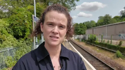 Anne Browne gives a TV interview on a train station platform. She has curly red hair and wears a collared black shirt.