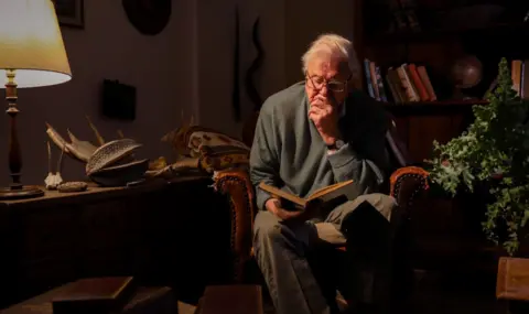 Toby Strong/Silverback Films/Netflix Sir David Attenborough sits on an armchair in a lowlit room and reads from a book. He wears a green jumper, slacks and glasses. A bookshelf is visible in the background. To his left is a house plant and a lamp can be seen on a table on his right. 