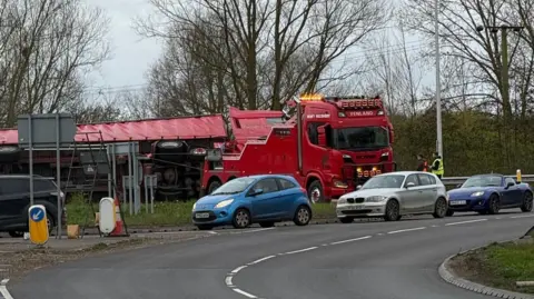 A red lorry broken down on the edge of a roundabout. Two men can be seen standing in the road at the front of the lorry looking at it. In front of the roundabout are queuing cars. 
