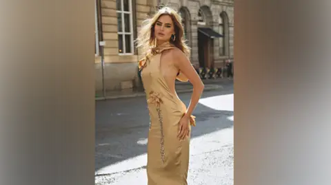 Peonies and Cream Macey wears the Leah dress while posing for the camera and standing in a street in Liverpool. The dress is sleeveless and a gold, yellow in colour with matching coloured flower decorations on with jewels for the stems. 