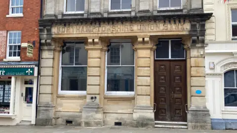 Empty stone-built building with two large windows and large wooden door.  The words "NORTHAMPTONSHIRE UNION BANK" can be seen in the stone above the windows and door. An adjacent estate agent is just visible to the left.