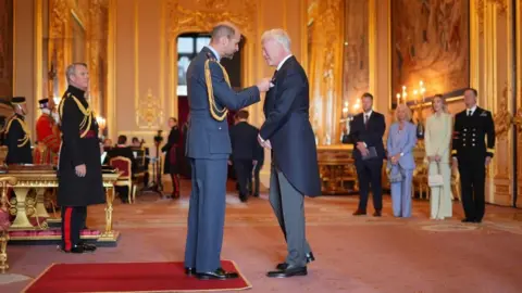 PA Media Prince William pins a medal onto the jacket of David Moyes. They are standing inside Windsor Castle. Other men in royal dress stand in the room. 