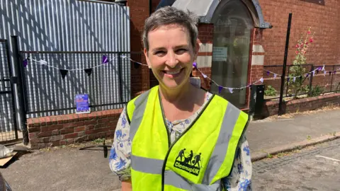 A smiling woman in a high-vis vest stands in the road with purple bunting behind her.