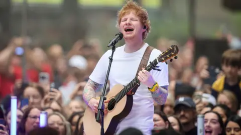 Reuters Ed Sheeran appears playing guitar, singing into a microphone, with people cheering behind him