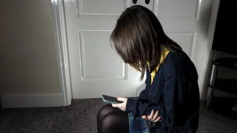 PA Media A dark-haired girl with her face hidden from view in school uniform sits cradling her arm whilst looking down at her mobile phone.