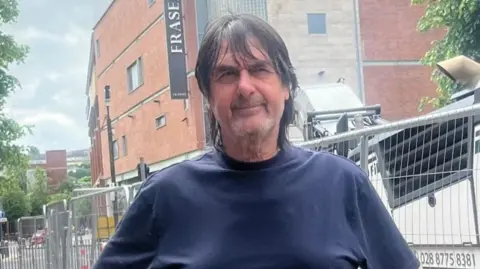 Record store owner lee Mason - a man with longish dark hair wear a dark blue tshirt. he is standing in a street in front of security barriers, with a large shopping centre in the background