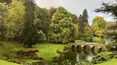A stone bridge crosses a lake. It is flanked either side by rolling lawns and arboretum trees.