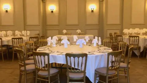 Tables decorated with napkins and cutlery in a large cream paneled function room with round tables 