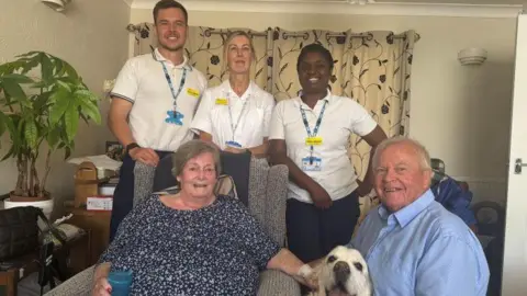 Charlotte North A woman in a flowery blue top and a man in a blue shirt, with a dog and three people in doctors uniform.