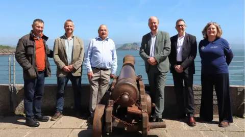 Six people stood next to a cannon (three either side). In the background is the sea and a small island (Drakes Island). The sky is blue. 