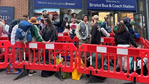 Josh Perkins/BBC A group of people queue between orange barriers for the bus from Bristol Bus Station to the Glastonbury Festival. They are wearing waterproofs and hats and many are carrying large rucksacks.