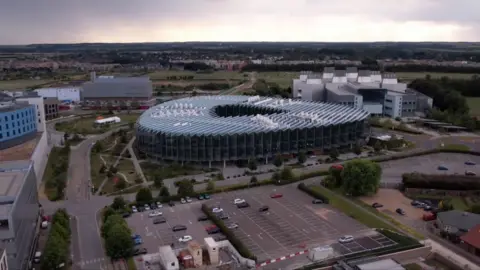 An aerial view of AstraZeneca's headquarters. In the centre of the picture is a modern oval-shaped building, which is covered in glass and whose roof is rippled and angular. A surface car park is in the foreground of the picture, while behind the large building in the centre are other modern-looking buildings, some housing in the nearer distance, then countryside. 