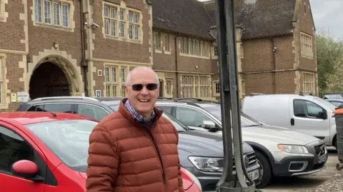 Martin Heath/BBC Martin Johns with short white hair and glasses smiling at the camera and wearing a brown bomber jacket. He is standing in front of a line of cars which a two-storey 1940s building behind him.