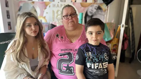 A mother wearing a pink printed t-shirt sits between her two children, an 18 year old wearing a white shirt and a 6 year old wearing a black and blue FIFA top