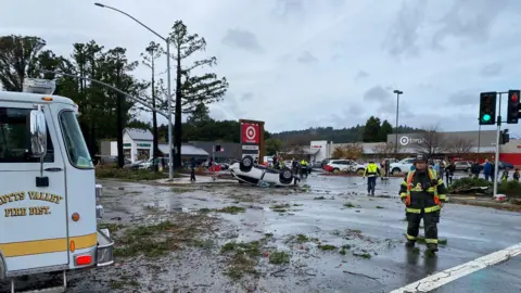 Scotts Valley Police Department A small white car flipped over in front of a large shop and car park in Scotts Valley. A fire truck and fireman can be seen in the foreground standing alongside debris from nearby trees. 