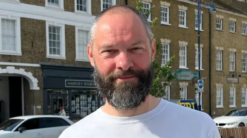 Tom Jackson/BBC Richard Slade is standing in the square while wearing a white top. He is smiling at the camera and has short hair and a beard, both are a mix of brown with some grey. Behind him is some seating on the square and trees and three-storey townhouse buildings.