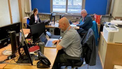 In a ground floor office, two men and a young woman sit at desks, each with black computer keyboards and monitors in front of them. 