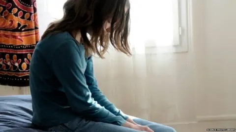 Science Photo Library A young girl sitting on a bed, looking sad