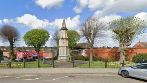 Google Stone war memorial with two trees on either side and well-kept gardens behind