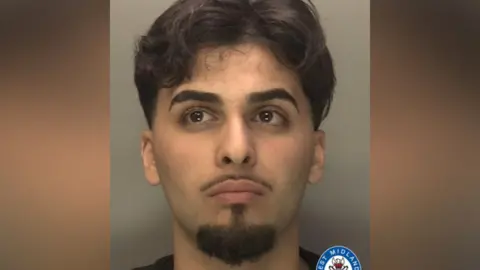 Deara Sadiq has long black hair and black beard, goatee and moustache. He is wearing a black top.