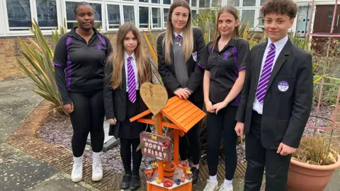 Shariqua Ahmed/BBC Poppi's friends, four girls in black and white school uniform and a boy, also in uniform, standing in front of an orange wooden memorial with clay poppies. The memorial is in a garden. It is surrounded by school buildings.