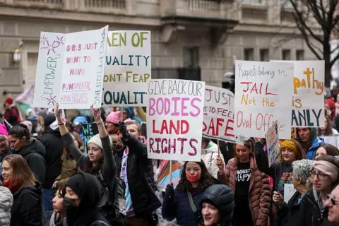 Getty Images Protesters hold signs. One reads "Give back out bodies land rights" 