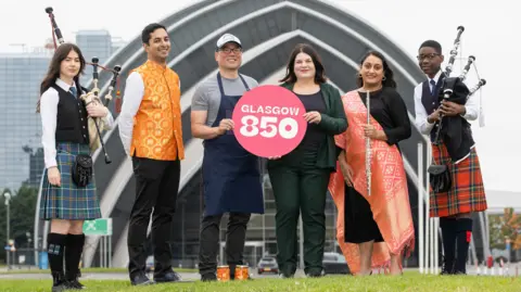 Glasgow 850 programme launch in front of the Armadillo venue on the north bank of the Clyde. Left to right: Sasha representing Piping Live! wearing a blue kilt and holding bagpipes, Sanjay from Glasgow Mela wearing an orange Modi waistcoat, Eddie Kim and council leader Susan Aitken holding a Glasgow 850 badge, Diljeet from the Mela holding a flute, and Kaylan in a red kilt with bagpipes.