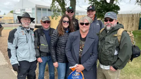 A group of race fans in leather jackets, puffa jackets, sunglasses and caps stand together near some trees and a marquee.